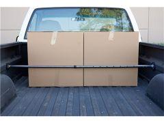 A pickup truck bed with two upright cardboard boxes secured by a horizontal black metal bar, set against a textured black surface.