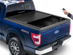 Rear view of a blue Ford F-150 pickup truck with a partially retracted black hard cover on the truck bed, and a person holding a remote control.