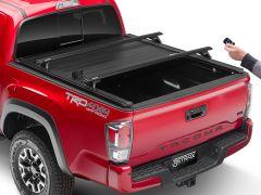 Rear view of a red Toyota Tacoma truck with a retractable tonneau cover, featuring the "TACOMA" name on the tailgate and a hand holding a remote control.