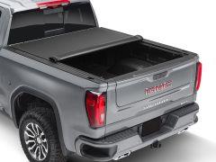 Rear view of a gray GMC Sierra pickup truck with a rolled-up black tonneau cover and rugged tires.
