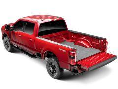 Red full-size pickup truck shown from rear three-quarter view with the tailgate lowered, revealing a gray carpeted bed mat installed in the truck bed; black step bars, rugged tires, and clean studio background visible.