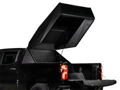 A close-up of a black pickup truck against a white background with the tailgate down and a large, angular truck bed topper lid raised, revealing the open cargo area and interior storage paneling.