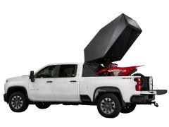 A white pickup truck with an open tailgate is shown from the side. In the truck bed, a black angular storage box is lifted open, revealing a red dirt bike secured inside.
