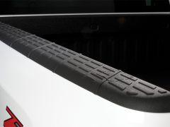 Close-up of a textured black rubber edge on a white truck bed, showcasing ridges and grooves for grip. A red emblem is partially visible.